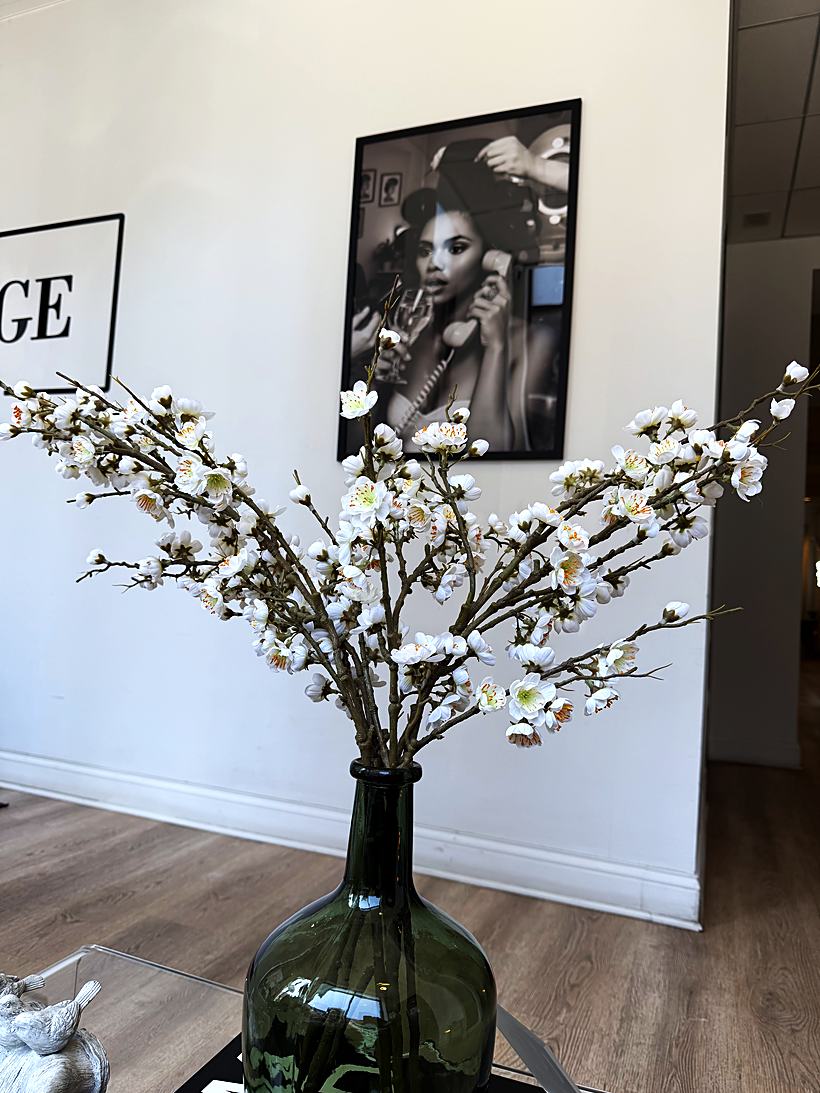 White blossoms in a green glass vase in front of framed salon artwork